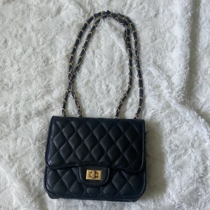Svart quiltad axelväska med kedja - Säljer en svart quiltad axelväska med guldfärgad kedja och snyggt vridlås framtill. Väskan har klassisk rektangulär form, innerfack med dragkedja och detaljer i guld. Perfekt för dig som gillar stilrena accessoarer med lyxig känsla.