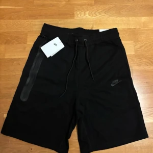 Svarta Nike shorts M - Svarta shorts från Nike i storlek M med elastisk midja och snörning. Snyggt minimalistisk design med diskret Nike-logga på benet och en svart tejpad ficka på sidan. Perfekta för träning eller chill dagar. Mjukt material som känns skönt mot huden.