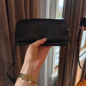 Svart väska från Zara - En superfin lite mindre väska från Zara. Den har två fack och ett extra fack som likar en korthållare. Den är försluten med en knapp. Väskan är i svart med guldiga detaljer, med följer en axelrem. Väskan är även snygg att hålla i handen. Hör av dig vid frågor💗☺️