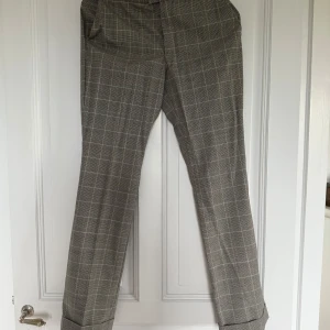 Rutig kostymbyxa i ullmix - Snygga grå kostymbyxor med rutig mönster och klassisk rak passform. Byxorna har uppvikta benslut och är tillverkade i en mjuk ullmix med polyester, elastan och en touch av lurex för extra känsla. Perfekta för en stilren och trendig look.