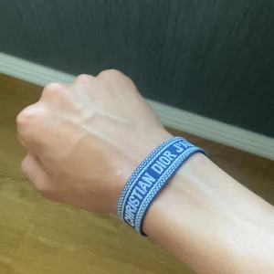 Blått Christian Dior armband - Snyggt blått armband från Christian Dior med broderad logga i vitt. Armbandet är i tyg och har knytning med tofsar i ändarna, vilket ger en chill och trendig vibe. Perfekt accessoar för att lyfta din outfit med lite lyxig känsla.