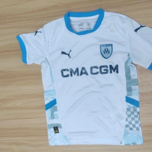 Marseille fotbollströja Puma vit/blå - Snygg Marseille fotbollströja från Puma i vitt med ljusblå detaljer och klubbmärke på bröstet. Tröjan har korta ärmar, blå krage och ärmslut samt tryckt sponsorlogga. Tillverkad i lätt och ventilerande material, perfekt för match eller träning.