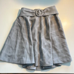 Grå rutig kortkjol med bälte - Grå kortkjol med rutigt mönster och stretchig midja med matchande bälte i tyg. Kjolen har en lätt utsvängd form och är fodrad med glansigt tyg på insidan. Köpt från okänd butik i Tokyo.