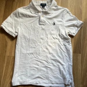 Vit pikétröja från Polo Ralph Lauren - Klassisk vit pikétröja från Polo Ralph Lauren i bomull. Tröjan har korta ärmar, krage med knappar och den ikoniska broderade loggan på bröstet. Perfekt för en clean och stilren look. Den är xl ungdom så passar s-m ungefär.                     Priset kan så klart diskuteras 