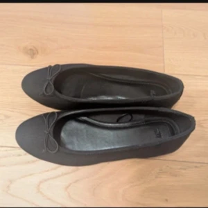 Svarta ballerinaskor med rosett - Stilrena svarta ballerinaskor med rund tå och söt rosettdetalj framtill. Skorna har en platt sula och är perfekta för en enkel och klassisk look. Materialet ser ut att vara syntet, vilket gör dem lätta att bära hela dagen.
