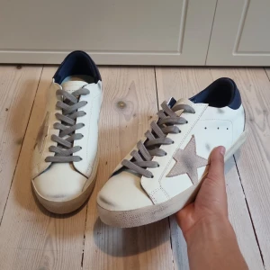 Golden Goose(pris kan diskutaras!) - Golden Goose sneakers i vitt med grå mockastjärna på sidan, grå snörning och mörkblå häl. Klassisk platt sula och rund tå. Snygg kontrast mellan färgerna och ikonisk stjärndetalj som ger en cool vibe. Hör av er om ni har en fråga eller vill diskutera priset