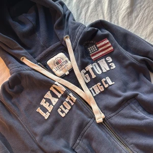 Marinblå hoodie från Lexington - Snygg marinblå hoodie från Lexington med broderad amerikansk flagga och stora vita tryck på bröstet. Tröjan har huva med vita snören, dragkedja framtill och känguruficka. 