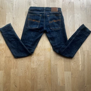 Mörkblå jeans från Nudie - Snygga mörkblå jeans från Nudie med klassisk orange söm på bakfickorna🙌. Modellen är skinny fit med normal midja och tillverkad i mjukt bomullsjeans. Perfekta för dig som gillar en smal siluett och stilren look. Storleken är w26/L32 Hör av er om ni har några frunderingar🤝kom med prisförslag så kan vi säkert komma överäns om något👏