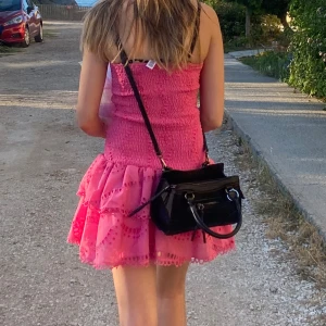 Rosa kort volangklänning - Supersöt rosa kortklänning med smockad överdel och volangig kjol. Klänningen är ärmlös och har fina broderade detaljer längs volangerna. Perfekt för varma dagar och ger en riktigt somrig vibe.