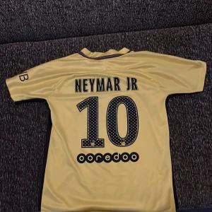 PSG Neymar Jr 10 gul fotbollströja - Gul Paris Saint-Germain fotbollströja med Neymar Jr och nummer 10 på ryggen. Tröjan har svarta detaljer, klubbmärke på bröstet och sponsorlogga framtill. Kortärmad modell i lätt och ventilerande material. Stl 146/152.