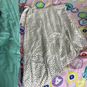 Prickig omlottkjol från Kappahl - Supersöt vit omlottkjol från Kappahl med svarta prickar och knyt i midjan. Kjolen har volangkant och är gjord i mjuk bomull, perfekt för en chill och trendig look. Passar dig som gillar lekfulla och klassiska mönster.