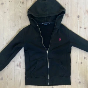 Svart zip up hoodie från Ralph Lauren  - Svart hoodie från Ralph Lauren Sport i 100% bomull. Klassisk modell med dragkedja, huva och kängurufickor. På bröstet finns den ikoniska rosa broderade polospelaren. Perfekt för en chill och sportig look.
