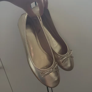 Guldiga ballerinaskor från H&M - Snygga ballerinaskor i guld från H&M med rund tå och söt rosettdetalj framtill. Skorna har en klassisk, platt sula och är tillverkade i syntetmaterial med glansig finish. Jättesnygga till fest!
