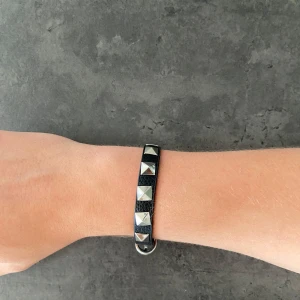Valentino liknande armband  - Coolt svart armband i läderimitation med fyrkantiga silverfärgade nitar längs hela bandet. Justerbart spänne i silverfärg gör att det passar de flesta handleder. Perfekt accessoar för en edgy look.