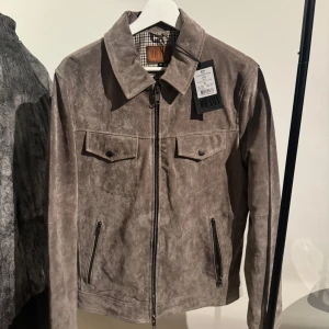 Brun mockajacka från Jofama - Snygg brun mockajacka från Jofama med klassisk krage, dragkedja framtill och två bröstfickor med lock. Jackan har raka linjer och diskreta sömmar, perfekt för dig som gillar stilren design. Fodret har rutigt mönster för extra detalj.