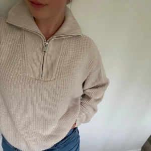 Beige stickad half zip tröja - Supermysig beige stickad tröja med half zip och hög krage. Tröjan har en relaxed passform och långa ärmar, perfekt för lager på lager. Ribbstickad struktur som ger en snygg och enkel look. Passar till jeans eller kjol för en chill vibe.