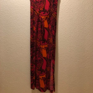 Färgstark långklänning från Indiska - Säljer en maxiklänning från Indiska med färgstarkt mönster i rött, orange och rosa. Klänningen är ärmlös, har rund halsringning och ett coolt paisleyinspirerat tryck. Perfekt för dig som vill sticka ut med en bohemisk vibe.