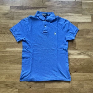 Ralph Lauren piké - Klassisk blå pikétröja från Polo Ralph Lauren med korta ärmar, krage och två knappar framtill. Ikoniska gula loggan broderad på bröstet. Tillverkad i mjuk bomull som andas och har en normal passform. Perfekt för en clean och sportig look. Jag skickar ej bild när jag bär plaggen och jag vet ej vilken längd och vikt de passar då jag inte prövar kläderna jag säljer 😃