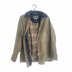 Barbour trenchcoat  - Klassisk brun parkajacka från Barbour med rutigt innerfoder och krage i manchester. Jackan har dragkedja och knappar framtill samt flera praktiska fickor. Perfekt för dig som gillar tidlös brittisk stil.