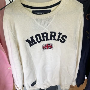 Vit stickad tröja från Morris - Vit stickad tröja från Morris med broderad logga och Union Jack-flagga på bröstet. Tröjan har rund halsringning, ribbade muddar och en klassisk, avslappnad passform. Perfekt för dig som gillar preppy stil och snygga detaljer.