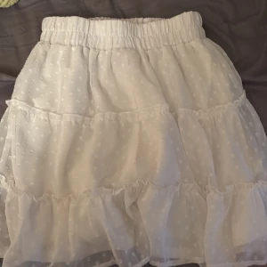 Vit volangkjol från FB Sister XS - Supersöt vit kortkjol från FB Sister i storlek XS. Kjolen har flera lager med volanger och ett prickigt mönster i tyget. Resår i midjan för skön passform och ett tunt, lätt material som känns luftigt. Perfekt för en fräsch och somrig look!