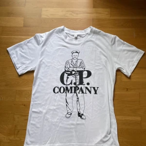 Vit C.P. Company t-shirt med tryck  - Vit t-shirt från C.P. Company med stort svart tryck framtill som föreställer en person och loggan. Klassisk rund hals och korta ärmar. Perfekt för dig som gillar streetwear och vill sticka ut med en ikonisk design.