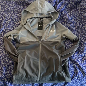 Grå vindjacka från Under Armour S - Snygg grå vindjacka från Under Armour med huva och dragkedja hela vägen. Jackan har en stilren look med logga på bröstet och praktiska fickor. Perfekt för dig som vill ha en sportig och clean stil. Materialet är lätt och känns smidigt.