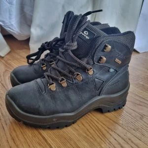 Svarta kängor från Graninge - Robusta svarta kängor från Graninge med grov sula och snörning. Tillverkade i slitstarkt skinn och med detaljer i metall. Perfekta för utomhusaktiviteter och äventyr. Svensk flagga på sidan och vadderad krage för extra komfort.