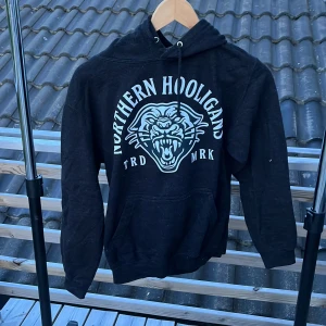 Svart hoodie Northern Hooligans XS - Svart hoodie från Northern Hooligans i storlek XS. Stor vit tryckt logga och pantermotiv på bröstet, klassisk huva med snörning och känguruficka framtill. Perfekt för dig som gillar streetwear och vill sticka ut.