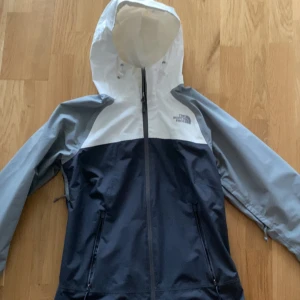 The North Face vindjacka blå/vit - Snygg vindjacka från The North Face i blått, vitt och grått. Jackan har huva med snörning, hel dragkedja framtill och två fickor med dragkedja. Perfekt för blåsiga dagar och outdoor-äventyr.