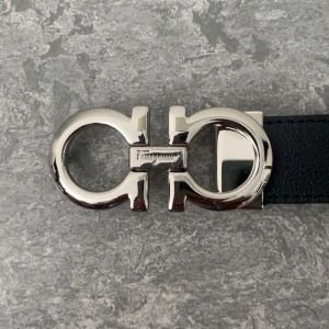 Svart Ferragamo skärp med silverdetalj - Snyggt svart bälte från Ferragamo med ikoniskt dubbel-Gancini-spänne i blankt silver. Bältet är gjort i äkta skinn och har en stilren, klassisk design som lyfter vilken outfit som helst. Perfekt för dig som gillar exklusiva accessoarer och vill sticka ut.