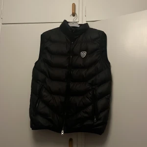 EA7 väst - Snygg svart dunväst från EA7 Emporio Armani med hög krage och två dragkedjefickor. Västen har ett stilrent EA7-märke på bröstet och quiltad design. storlek S-M 