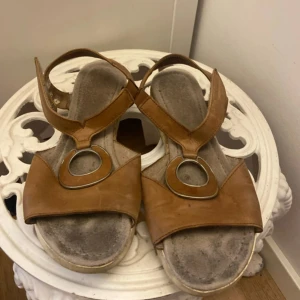 Bruna sandaler från Gabor med ringdetalj - Snygga bruna sandaler från Gabor i skinn med breda remmar och cool metallring framtill. Bekväm fotbädd och platt sula, perfekt för varma dagar. Klassisk design med modern twist, passar till många outfits.