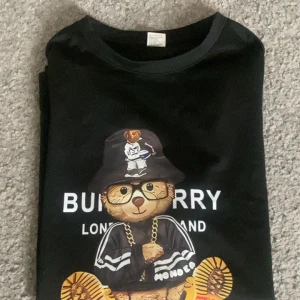 Svart t-shirt med tryck Burberry - Svart t-shirt från Burberry med stort färgglatt tryck framtill som visar en tecknad figur med keps, guldkedja och sneakers. Klassisk rund halsringning och korta ärmar. Snygg streetstyle-vibe och cool design.