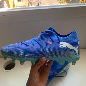 Blåa Puma fotbollsskor strl 39 - Snygga blåa fotbollsskor från Puma med vita detaljer och logga på sidan. Skorna har snörning, mönstrad ovandel i syntetmaterial och ljusblå dobbar för bra grepp på planen. Perfekta för dig som vill sticka ut på matchen! OBS box ingår inte.