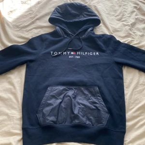  Tommy Hilfiger huvtröja  - Snygg mörkblå hoodie från Tommy Hilfiger med broderad logga och text på bröstet. Tröjan har huva med snörning och en stor ficka framtill i glansigt material. Perfekt för dig som gillar klassisk streetstyle och vill ha något bekvämt och stilrent.