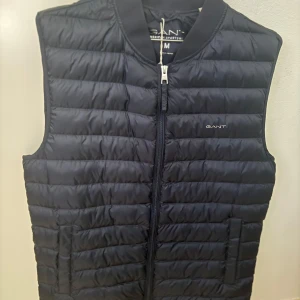 Svart dunväst från GANT - Snygg svart dunväst från GANT med quiltad design och dragkedja framtill. Västen har diskret GANT-logga på bröstet och nacken, samt två fickor på sidorna. Perfekt att slänga över en hoodie eller tröja för extra värme.