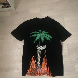 Palm Angels Firestarter t-shirt svart - Svart t-shirt från Palm Angels med stor palm, skelett och eld på framsidan. Baksidan har vit text med 'FIRESTARTER' och Palm Angels-logga. Skön passform och coolt tryck som sticker ut. Perfekt för dig som gillar streetwear och edgy stil.