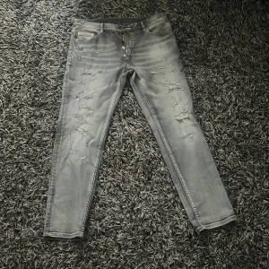 Grå slitna jeans från Dsquared2 - Säljer ett par grå jeans från Dsquared2 med coola slitningar och tvättad look. Byxorna har fem fickor, dragkedja och smal passform. Märkesdetalj bak med '1964'. Perfekta för dig som gillar streetstyle och vill sticka ut.