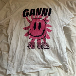 Vit Ganni t-shirt med rosa smiley - Cool vit t-shirt från Ganni med stor rosa smiley och svart text framtill. Oversized fit och klassisk rund halsringning. Perfekt statement-plagg med unik grafisk print. Tillverkad i mjuk bomull som känns skön mot huden.