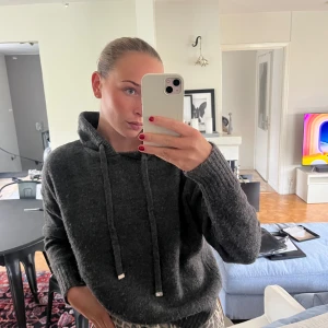 Mörkgrå stickad hoodie med huva - Mysig mörkgrå stickad hoodie med huva och långa ärmar. Tröjan har breda ribbade muddar och snörning i halsen. Perfekt för kyliga dagar när du vill vara både bekväm och snygg.