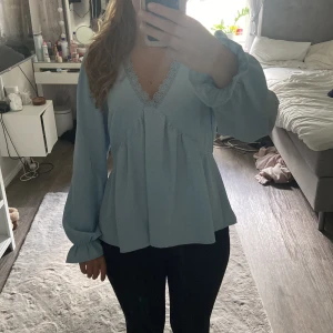 Ljusblå blus med spets från Vero Moda - Supersöt ljusblå blus från Vero Moda med v-ringning och spetskant. Blusen har långa puffiga ärmar med resår vid handlederna och en lös, flowy passform. Perfekt för dig som gillar romantiska och luftiga plagg.
