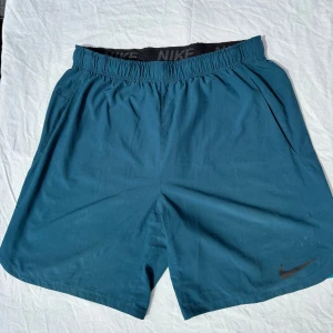 Blå Nike träningsshorts - Nike träningsshorts i marinblått med elastisk midja och svarta Nike-loggan på benet. Shortsen har fickor på sidorna och ventilerande mesh-detaljer i ljusare blått nedtill. Perfekta för sport eller gym och riktigt sköna att röra sig i.