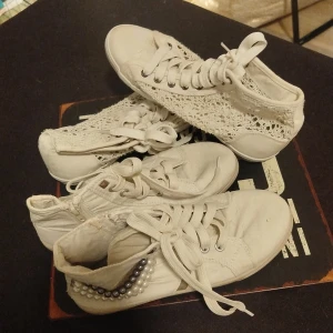 Två par  Vita höga sneakers med pärlor & spets. - Två par vita höga sneakers, ett par med coola pärldetaljer i vitt och grått på hälen och ett par med snygga virkade spetsdetaljer på sidorna. Båda har snörning och platt sula, perfekta för dig som vill sticka ut med din stil. Nya men sen tvättade i maskin då det var lite lim fläckar längs sulan som däremot inte gick bort. Inget som stör alls om jag ska säga min åsikt 😁 