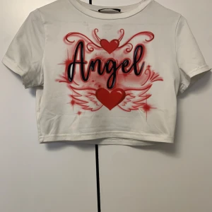 Vit croppad topp med Angel-tryck - Vit croppad topp med korta ärmar och stort airbrush-tryck framtill med texten 'Angel', röda hjärtan och vingar. Perfekt statement-plagg för dig som gillar unika och trendiga prints. Mjuk och skön bomullskänsla.