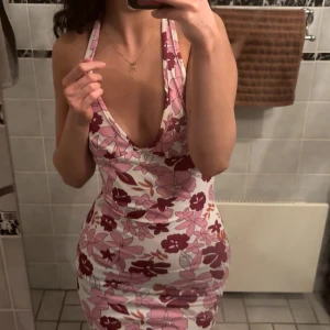Blommig kortklänning med djup urringning - Säljer en tight kortklänning med blommigt mönster i rosa, vinrött och vitt. Klänningen har djup v-ringning, öppen rygg och knyt i nacken. Perfekt för dig som vill sticka ut med en färgglad och somrig look. Passar xs-m