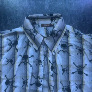 Randig skjorta med dödskallemönster - Säljer en kortärmad skjorta från Iron Cat i vitt och grått med gula ränder och svarta dödskallar över hela tyget. Skjortan har klassisk krage och knäppning framtill. Perfekt för dig som vill sticka ut med en edgy stil.