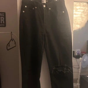 Svarta jeans från 157 med hål på knät - Svarta jeans från 157 med klassisk femficksmodell och knäppning med knappar. Jeansen har ett stort, slitet hål över ena knät för en edgy look. Materialet är jeans och passformen är rak. Perfekta för dig som gillar streetstyle.