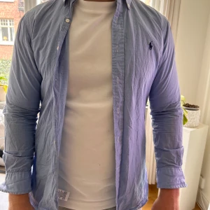 Rutig blå skjorta från Ralph Lauren - Snygg blå skjorta från Ralph Lauren i slim fit-modell. Skjortan har ett diskret rutmönster, klassisk krage och långa ärmar. Motsvarar en storlek M i min åsikt. Broderad logga på bröstet. Perfekt för dig som gillar stilrena och tidlösa plagg.