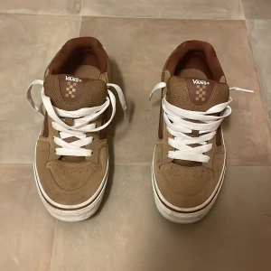 Bruna Vans sneakers med vit sula - Säljer ett par bruna Vans sneakers med vit sula och vita snören. Skorna har en klassisk låg profil och rund tå, med detaljer i mocka och syntet. Insidan är också brun och de har den ikoniska Vans-loggan på plösen och hälen. Perfekt för dig som gillar streetstyle.